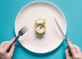 Hands holding knife and fork above alarm clock on a plate on blue background. Intermittent fasting, Ketogenic dieting, weight loss, meal plan, and healthy food concept.